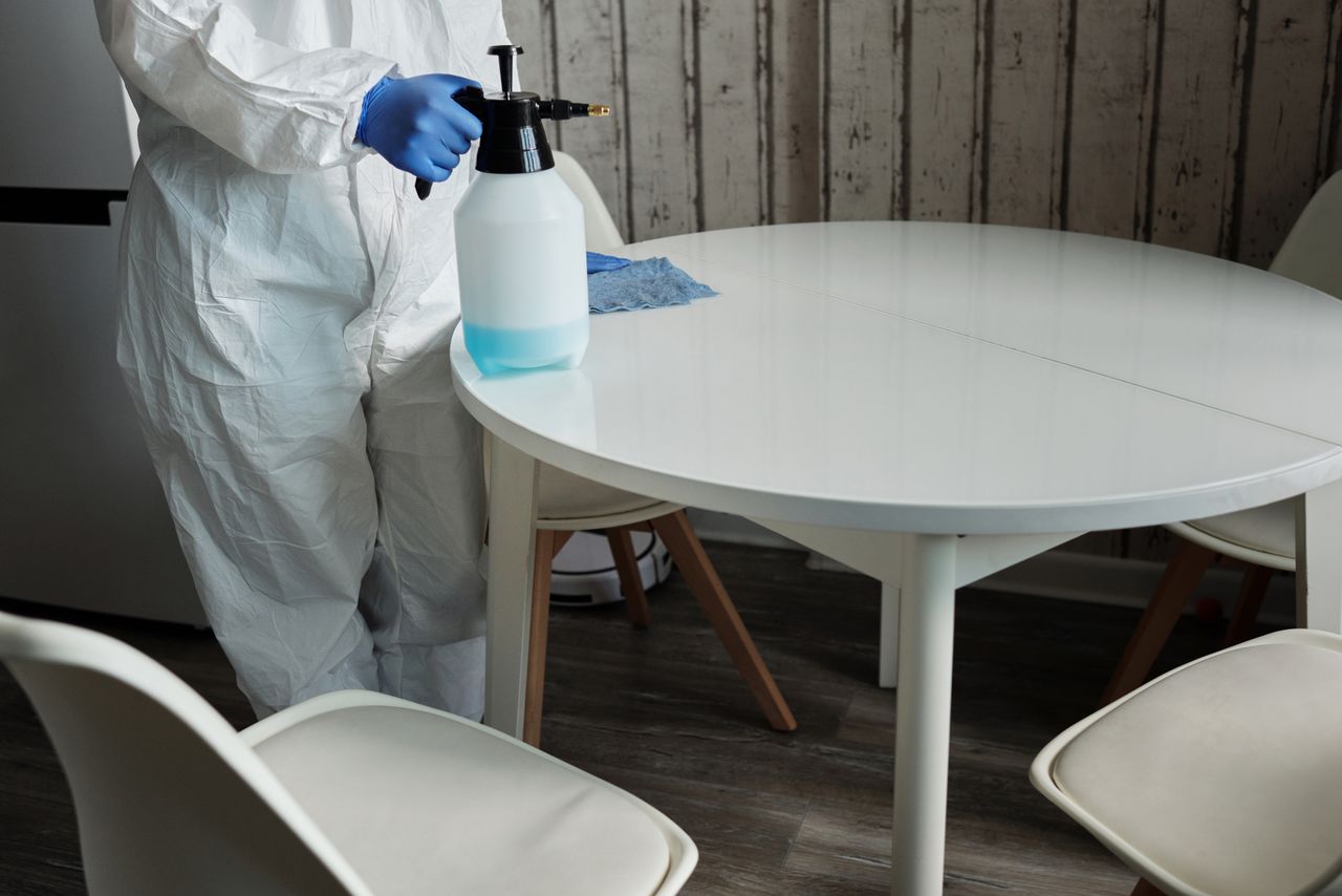 services-04 Person in protective suit disinfecting a white table indoors with a sprayer.
